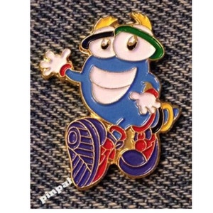 May include: A blue enamel pin depicting a cartoon character with a smiling face, wearing a hat with green and black accents. The pin has gold-coloured trim and red and white details.