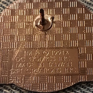 Op de afbeelding: Een gouden pin met een getextureerde achterkant. De achterkant van de pin heeft de tekst "TM & &copy; 1991x DC COMICS INC. MADE IN TAIWAN GIFT CREATIONS INC."