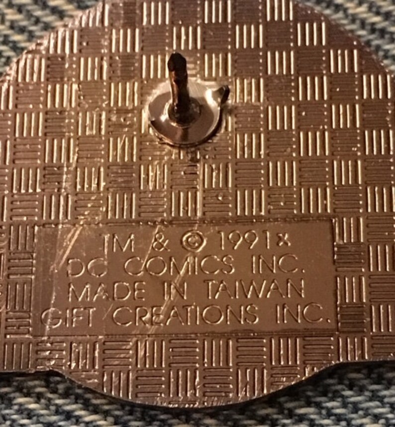 Op de afbeelding: Een gouden pin met een getextureerde achterkant. De achterkant van de pin heeft de tekst "TM & &copy; 1991x DC COMICS INC. MADE IN TAIWAN GIFT CREATIONS INC."