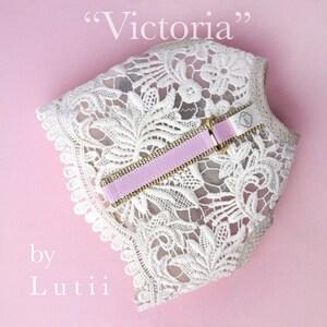 May include: White lace dog harness with a pink ribbon and gold buckle. The harness has a floral design and the text "Victoria" is written above it. The text "by Lutii" is written below the harness.
