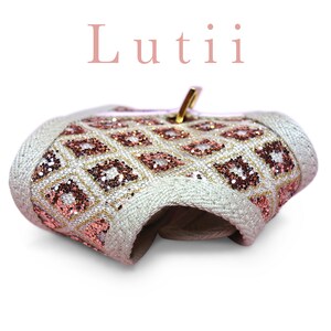 K&ouml;nnte beinhalten: Eine wei&szlig;e Stofftasche mit einem goldenen Rei&szlig;verschluss und einem geometrischen Muster in Pink und Gold. Die Tasche ist in der Mitte gefaltet und der Text "Lutii" ist &uuml;ber der Tasche sichtbar.