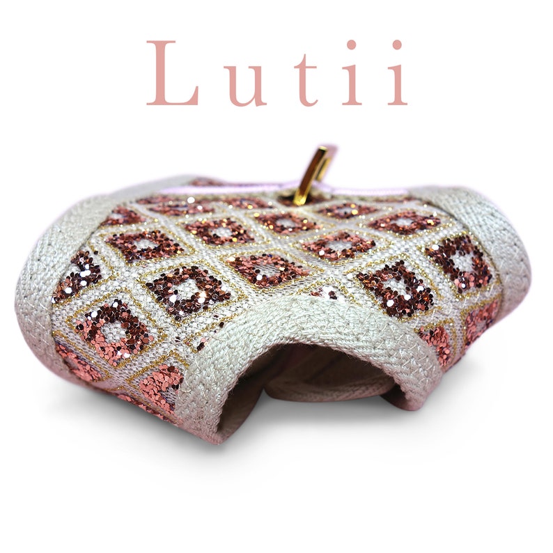 K&ouml;nnte beinhalten: Eine wei&szlig;e Stofftasche mit einem goldenen Rei&szlig;verschluss und einem geometrischen Muster in Pink und Gold. Die Tasche ist in der Mitte gefaltet und der Text "Lutii" ist &uuml;ber der Tasche sichtbar.