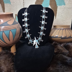 May include: A silver necklace with turquoise accents and a bird pendant is displayed on a black velvet bust. The necklace features a series of bird-shaped pendants and small silver beads. Two decorative pottery vases flank the necklace, set against a patterned textile background.