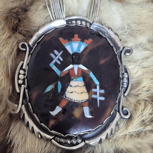 Puede incluir: Un collar con colgante de plata con un diseño central ovalado. La figura incrustada es roja, blanca y turquesa, con una cara negra y un tocado. El colgante está sobre un fondo marrón. El collar tiene una cadena de plata.