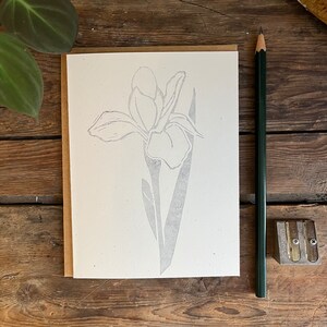 May include: A blank greeting card with a floral iris design in gray ink. The card is on a brown paper backing. A green pencil and pencil sharpener are next to the card. The card is on a wooden surface.