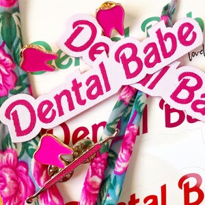 May include: Pink and white fabric patches with the text "Dental Babe" and a pink tooth pin. The patches are on a floral fabric background.