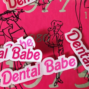May include: Five white fabric patches with pink lettering that reads "Dental Babe" and "The Babe". The patches are on a pink background with a black and white illustration of a woman.