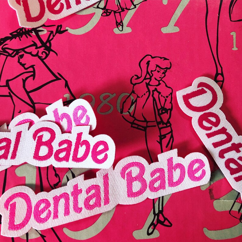 May include: Five white fabric patches with pink lettering that reads "Dental Babe" and "The Babe". The patches are on a pink background with a black and white illustration of a woman.