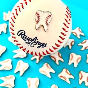 May include: A baseball with red stitching and a Rawlings logo, with a tooth-shaped pin. Scattered around are several tooth-shaped pins with baseball stitching. The background is a bright blue.