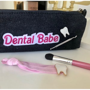 May include: A black denim zippered pouch with a white patch that says "Dental Babe" in pink letters. There are two white tooth-shaped pins, a pink makeup brush, and a pink hair tie.