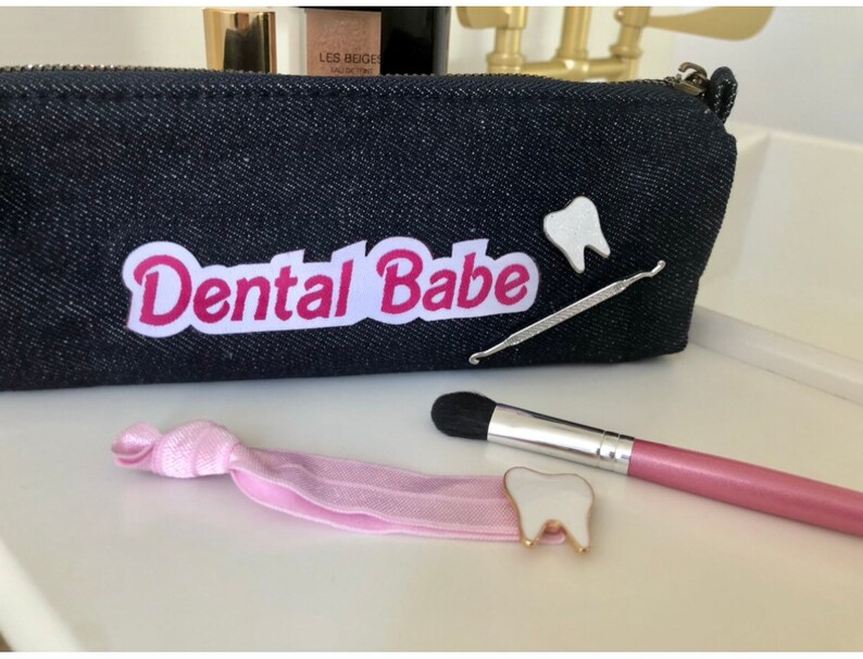 May include: A black denim zippered pouch with a white patch that says "Dental Babe" in pink letters. There are two white tooth-shaped pins, a pink makeup brush, and a pink hair tie.