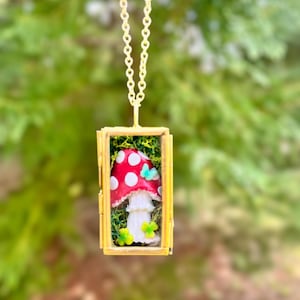 May include: A gold necklace with a rectangular locket featuring a red and white mushroom with white dots, green leaves, and a blue butterfly inside. The locket is surrounded by a gold frame.