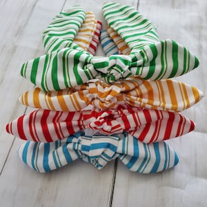 May include: A stack of four colorful striped headbands. The headbands are in green, yellow, red, and blue, each with white stripes. The headbands are knotted in a bow shape.