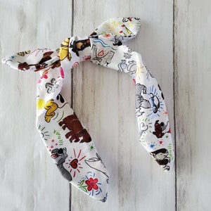 May include: White fabric headband with a colorful cartoon character print. The headband is tied in a bow and features various cartoon characters in brown, yellow, and gray, with red, blue, and green accents. The background is a light wood grain.