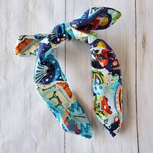 May include: A colorful headband with a space-themed pattern. The fabric features illustrations of rockets, planets, and stars in shades of blue, orange, and white. The headband is tied in a knot.