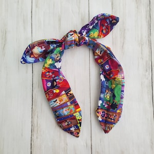 May include: A colorful, patterned hair bow featuring a repeating design of cartoon characters in various scenes. The bow is tied in a knot with two long, flowing ends. The background is a light wood grain.