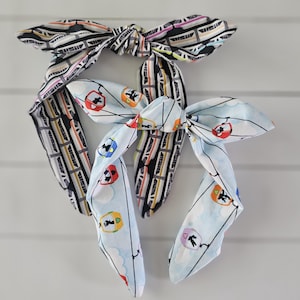 May include: Three fabric headbands with bow details. One headband features a black and white pattern with colorful accents. The other two headbands have a light blue background with a repeating graphic of a ski lift.
