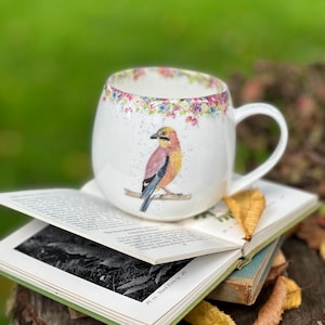 Puede incluir: Taza de cerámica blanca con una ilustración de un pájaro colorido, colocada sobre un libro abierto. La taza tiene un patrón floral en el borde y un asa. El fondo es verde borroso. La taza mide unos 10 cm de alto.