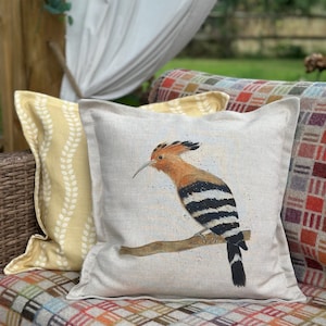 May include: Square throw pillow with a Hoopoe bird illustration. The bird has orange, black, and white feathers, perched on a brown branch. The pillow is on a patterned blanket with other pillows in the background.