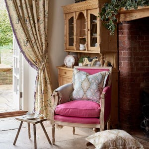 Pode incluir: Uma poltrona rosa com um almofada de padrão floral está em uma sala com um armário de madeira e uma lareira de tijolo. A poltrona está em frente a uma janela com vista para um jardim.