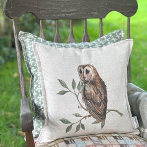 Puede incluir: Un cojín cuadrado decorativo con una ilustración detallada de un búho común posado en una rama con hojas verdes. El cojín es de color beige claro y descansa sobre una silla de madera con un cojín estampado verde detrás.