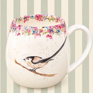 May include: A cream-coloured ceramic mug featuring a long-tailed tit bird perched on a brown branch. The rim is decorated with a colourful floral pattern of pink, blue, and yellow flowers. The mug has a speckled finish and a curved handle.