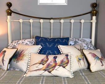 May include: A bed with a white metal frame and a green and blue plaid blanket. There are several decorative pillows with animal and nature designs, including a pheasant, a hedgehog, a deer, and a group of sheep.