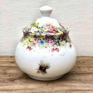Könnte beinhalten: Weiße Zuckerdose aus Keramik mit Blumenmuster und einer Biene auf der Vorderseite. Der Deckel hat ein Blumenmuster.