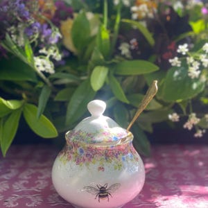 Puede incluir: Un azucarero de cerámica blanca con tapa y cuchara dorada. El cuenco está decorado con un estampado floral en rosa, azul y amarillo, y una ilustración de abeja negra y amarilla. El cuenco está sobre una superficie rosa.