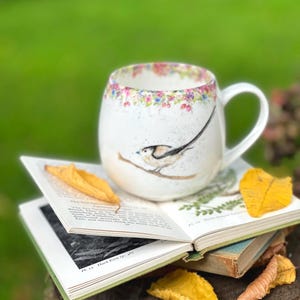 Op de afbeelding: Witte keramische mok met een vogelillustratie en een bloemenrand. De mok staat op een open boek, met herfstbladeren in de buurt. De achtergrond is een wazig groen. Een charmante mok met een natuurthema.
