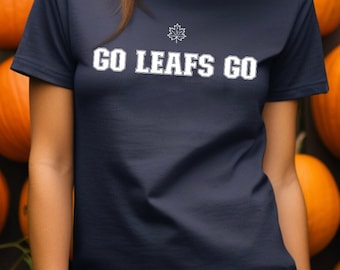 May include: Navy blue t-shirt with the text "GO LEAFS GO" in white. The shirt features a maple leaf logo above the text. The background includes a wall of orange pumpkins, suggesting a fall or autumn theme.