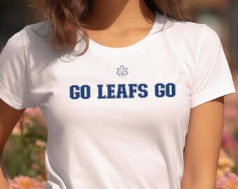 May include: White t-shirt with the text "GO LEAFS GO" in navy blue. The shirt features a stylized snowflake design above the text. The person is wearing white pants and is standing in front of a flower garden.