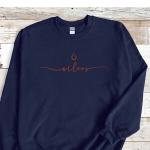 May include: Navy blue sweatshirt with the word "oilers" in a cursive font, and a flame graphic above the word. The text and graphic are in a burnt orange color. The sweatshirt is made of a soft material.