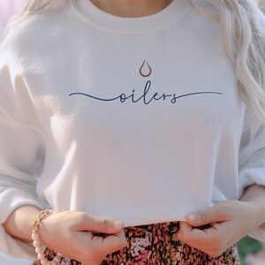 May include: White sweatshirt with the word "oilers" in a cursive font. Above the word is a stylized drop design in orange and navy blue. The sweatshirt is a crew neck style.
