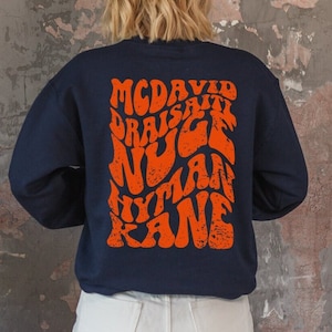 Może przedstawiać: Granatowa bluza z dużym pomarańczowym nadrukiem na plecach. Nadruk zawiera tekst "MCDAVID DRAISATI NUGE HYTLAN KANE" w retro, psychodelicznym stylu.