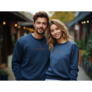 May include: Two people wearing navy blue sweatshirts. Each sweatshirt has a small orange flame graphic above the word "oilers" in a cursive font. The sweatshirts are a solid color and have a crew neck.