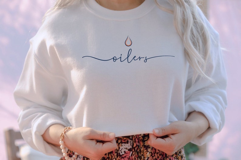 May include: White sweatshirt with the word "oilers" in a cursive font. Above the word is a stylized drop design in orange and navy blue. The sweatshirt is a crew neck style.
