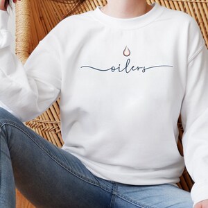 May include: White sweatshirt with the word "oilers" in a cursive font, with a small droplet graphic above the word. The sweatshirt has long sleeves and a crew neck. The overall style is casual and comfortable.