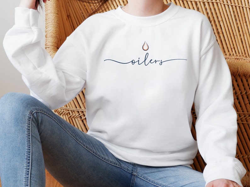 May include: White sweatshirt with the word "oilers" in a cursive font, with a small droplet graphic above the word. The sweatshirt has long sleeves and a crew neck. The overall style is casual and comfortable.