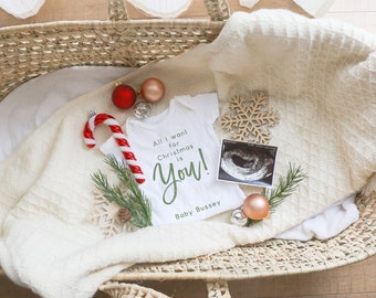 May include: A white onesie with the text "All I want for Christmas is You!" and the words "Baby Bussey" underneath. The onesie is in a wicker basket with a white blanket, a candy cane, a Christmas ornament, and a sonogram photo.