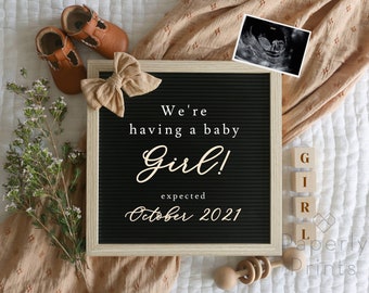 May include: A black letter board with the text "We're having a baby Girl! expected October 2021". The board is surrounded by a brown blanket, baby shoes, a bow, and a small wooden toy.