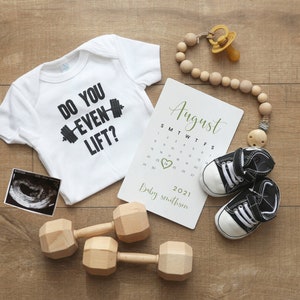 May include: A white onesie with the text "Do you even lift?" and a black dumbbell graphic. A calendar with the month of August highlighted, a pair of black and white baby shoes, a wooden baby teether, and a black and white ultrasound image.