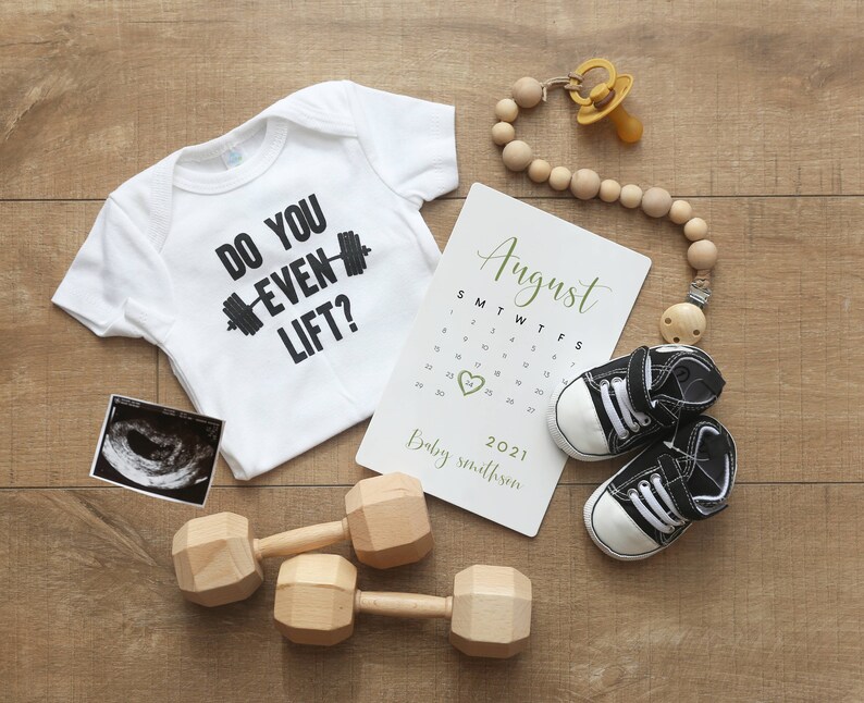 May include: A white onesie with the text "Do you even lift?" and a black dumbbell graphic. A calendar with the month of August highlighted, a pair of black and white baby shoes, a wooden baby teether, and a black and white ultrasound image.