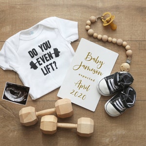 May include: A white onesie with the text "Do you even lift?" and a black dumbbell graphic. A white card with the text "Baby Jameson expected April 2020" is next to the onesie. There are two wooden dumbbells, a dummy, and a pair of baby shoes.