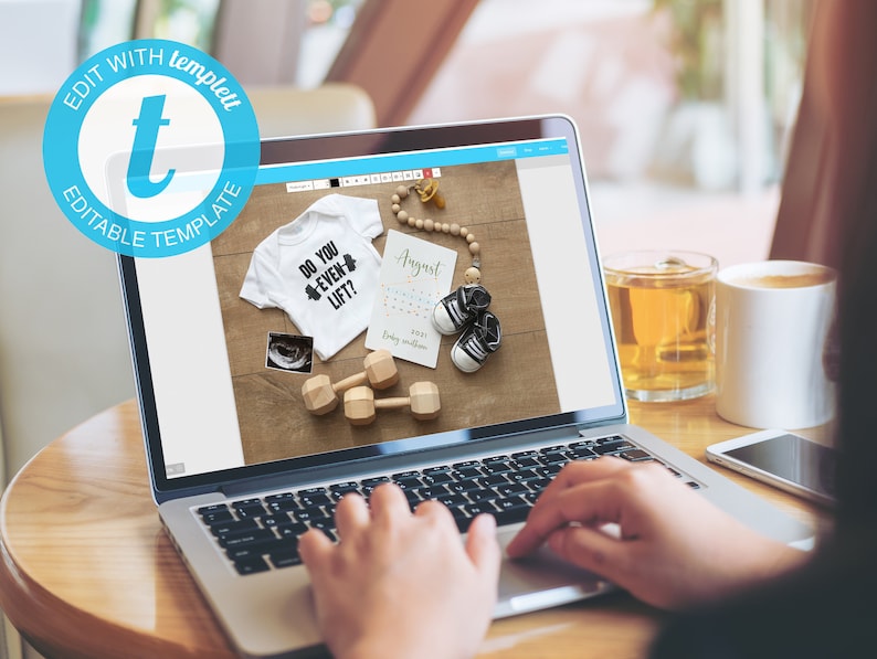 May include: A laptop computer displaying a website with a mockup of a baby onesie with the text "Do you even lift?" and a baby shoe. The onesie is white with black text and a black and white ultrasound image. The baby shoe is black with white trim. The website is on a light blue background with a white border. The laptop is on a wooden table.