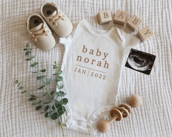 Puede incluir: Un body blanco para bebé con el texto "baby norah JAN 2022" impreso en él. El body está rodeado de bloques de madera que deletrean "BABY", un par de zapatos de bebé, un pequeño ramo de eucalipto, un sonajero de madera y una imagen de ultrasonido.