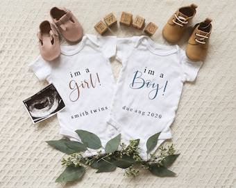 May include: Two white baby onesies with the text "I'm a girl!" and "I'm a boy!" written in cursive. The onesie with the girl text has the words "Smith Twins" below it. The onesie with the boy text has the words "Due Aug 2020" below it. There are two pairs of baby shoes, one pink and one brown, and a few wooden blocks with letters on them.
