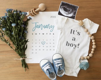 May include: A white onesie with the text "It's a boy!" in gray lettering. A calendar with the date January 12th circled in blue. A blue baby shoe, a wooden rattle, and a wooden bead necklace.