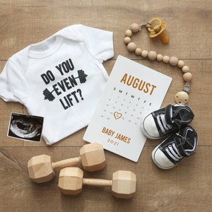 May include: A white babygrow with the text "Do you even lift?" and a black dumbbell graphic. A calendar with the month of August highlighted, a sonogram, two wooden dumbbells, and a wooden bead necklace with a dummy.