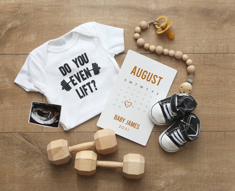 May include: A white babygrow with the text "Do you even lift?" and a black dumbbell graphic. A calendar with the month of August highlighted, a sonogram, two wooden dumbbells, and a wooden bead necklace with a dummy.